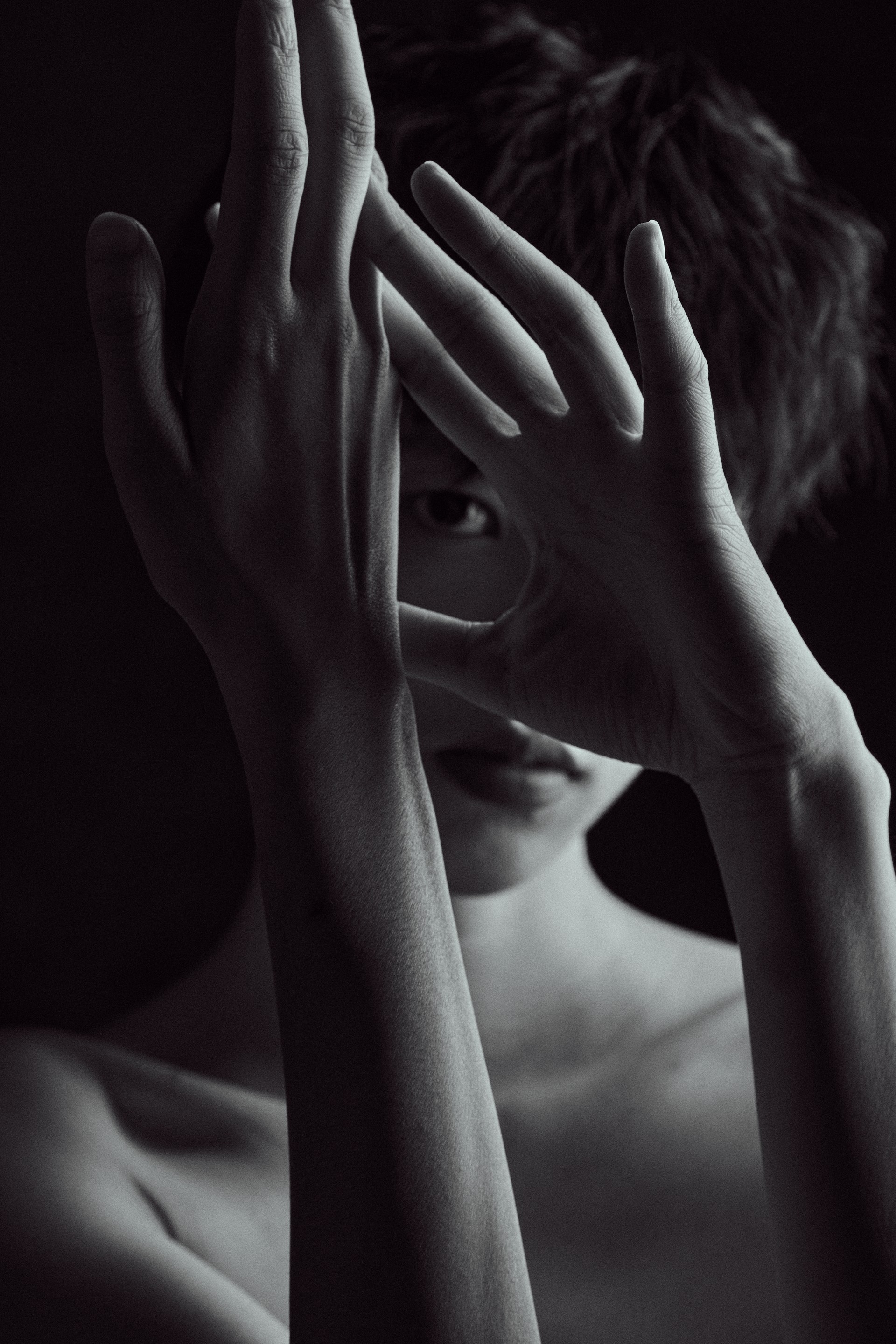 A black-and-white portrait with raised hands framing the eyes, photographed by Hill Xue