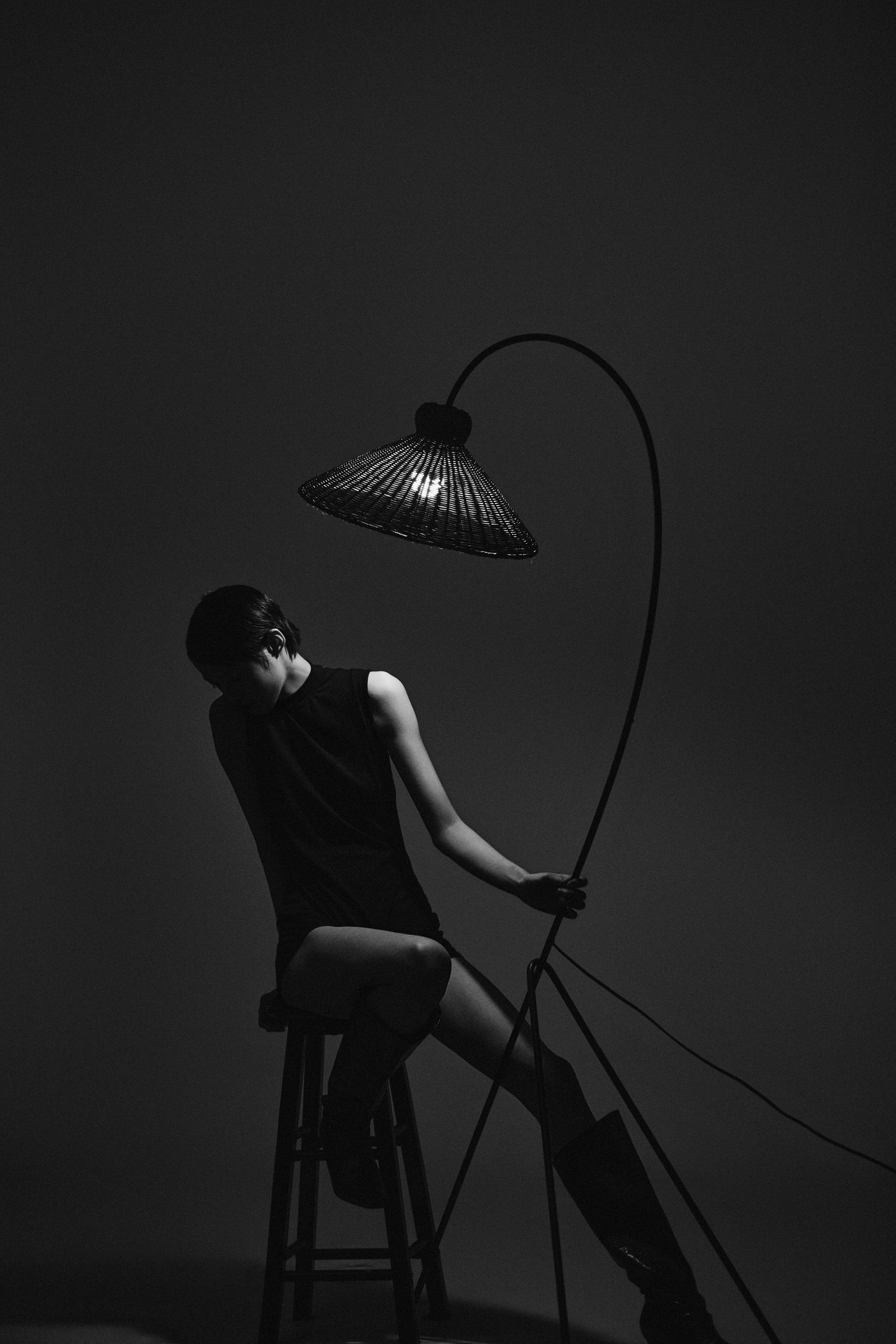 A black-and-white full-length seated portrait beside a curved floor lamp, photographed by Hill Xue