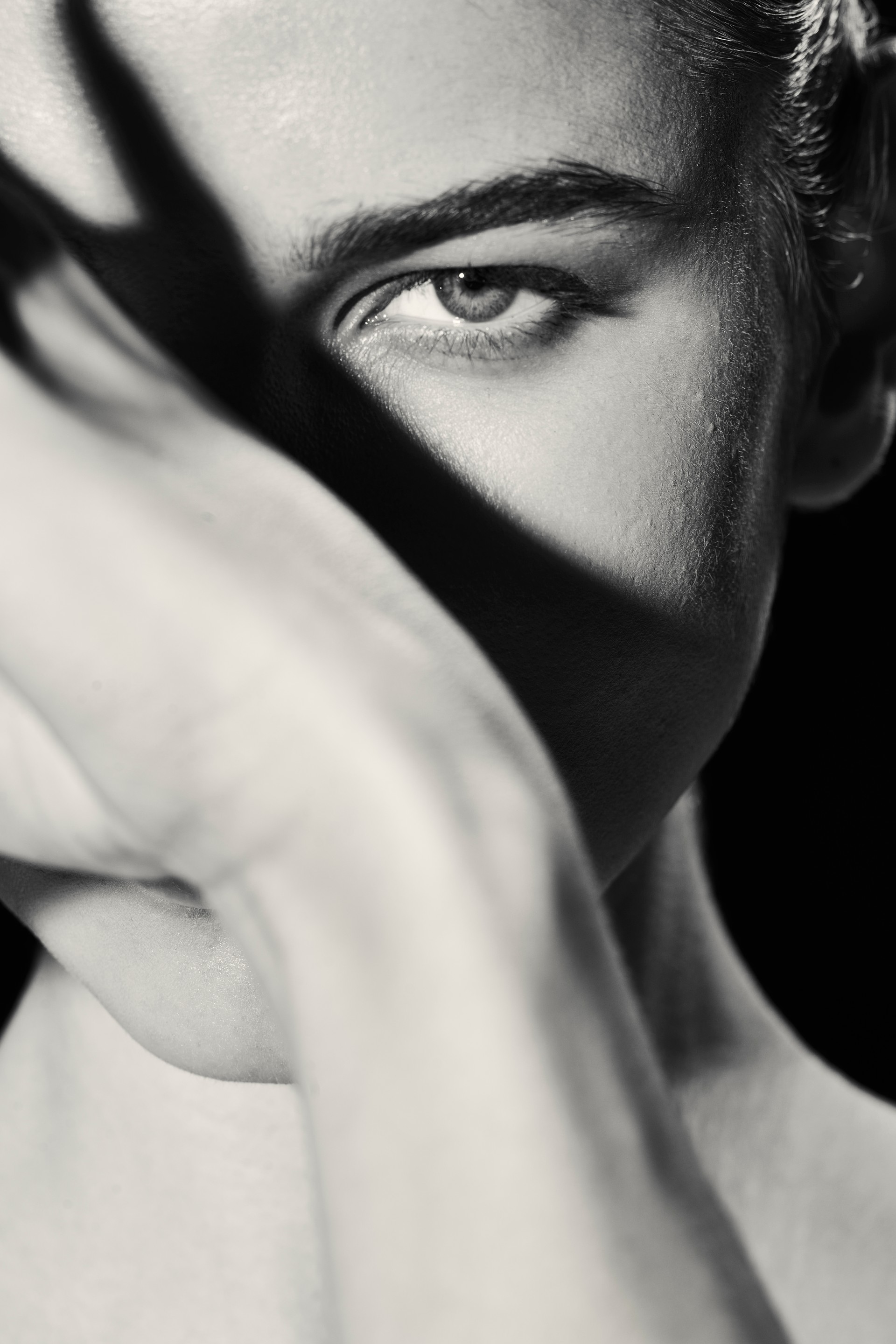 A black-and-white close-up portrait with one eye framed by a raised arm and dramatic shadows, photographed by Hill Xue