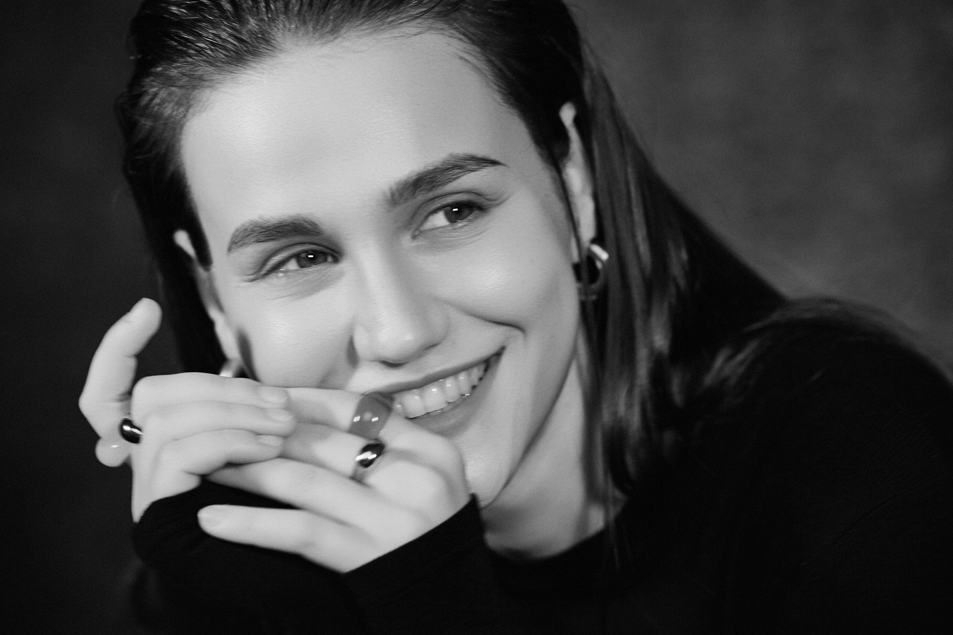 A smiling black-and-white close-up portrait with hands beneath the face, photographed by Hill Xue