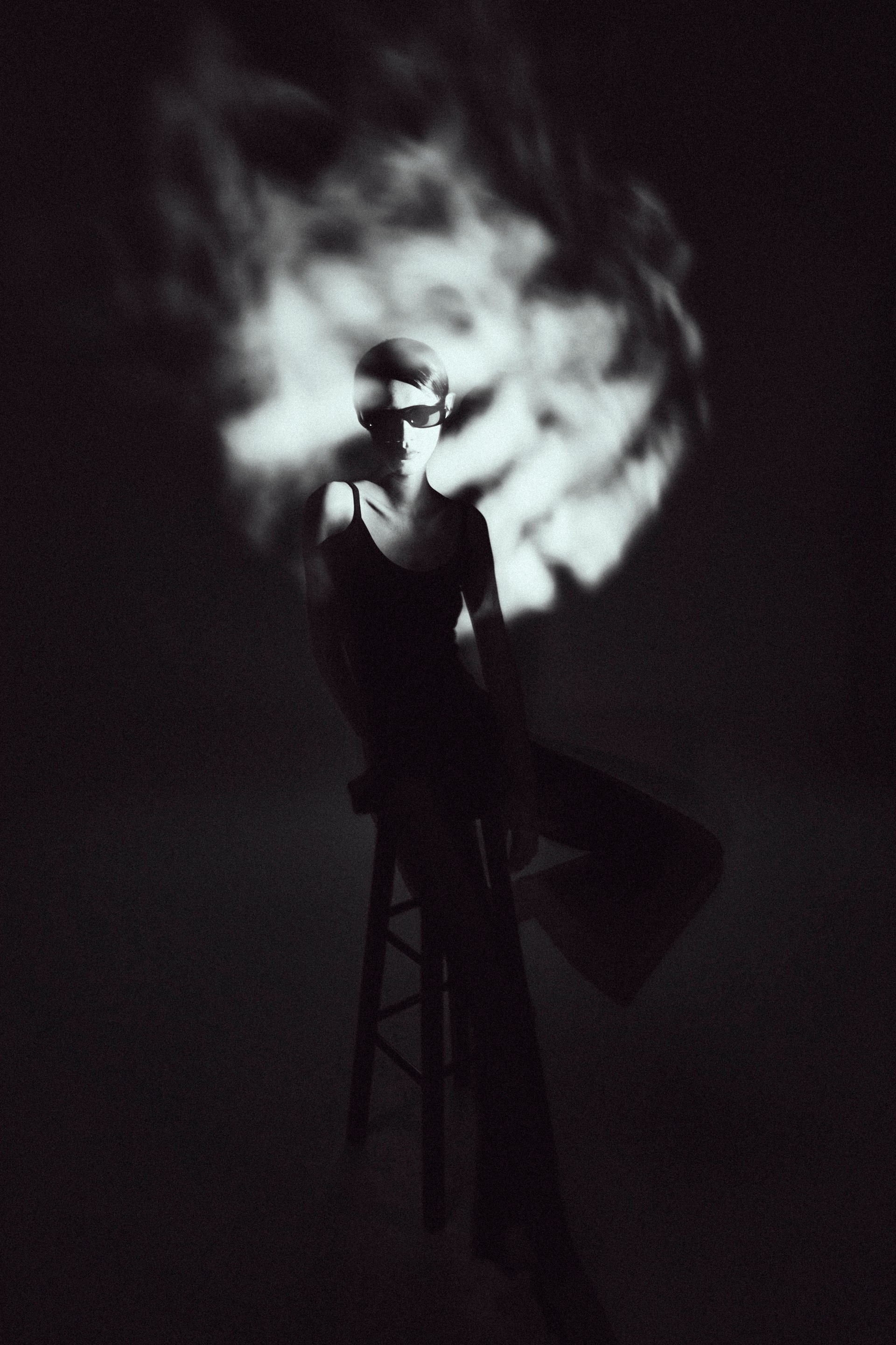 A seated black-and-white portrait wearing sunglasses against a patterned light projection, photographed by Hill Xue