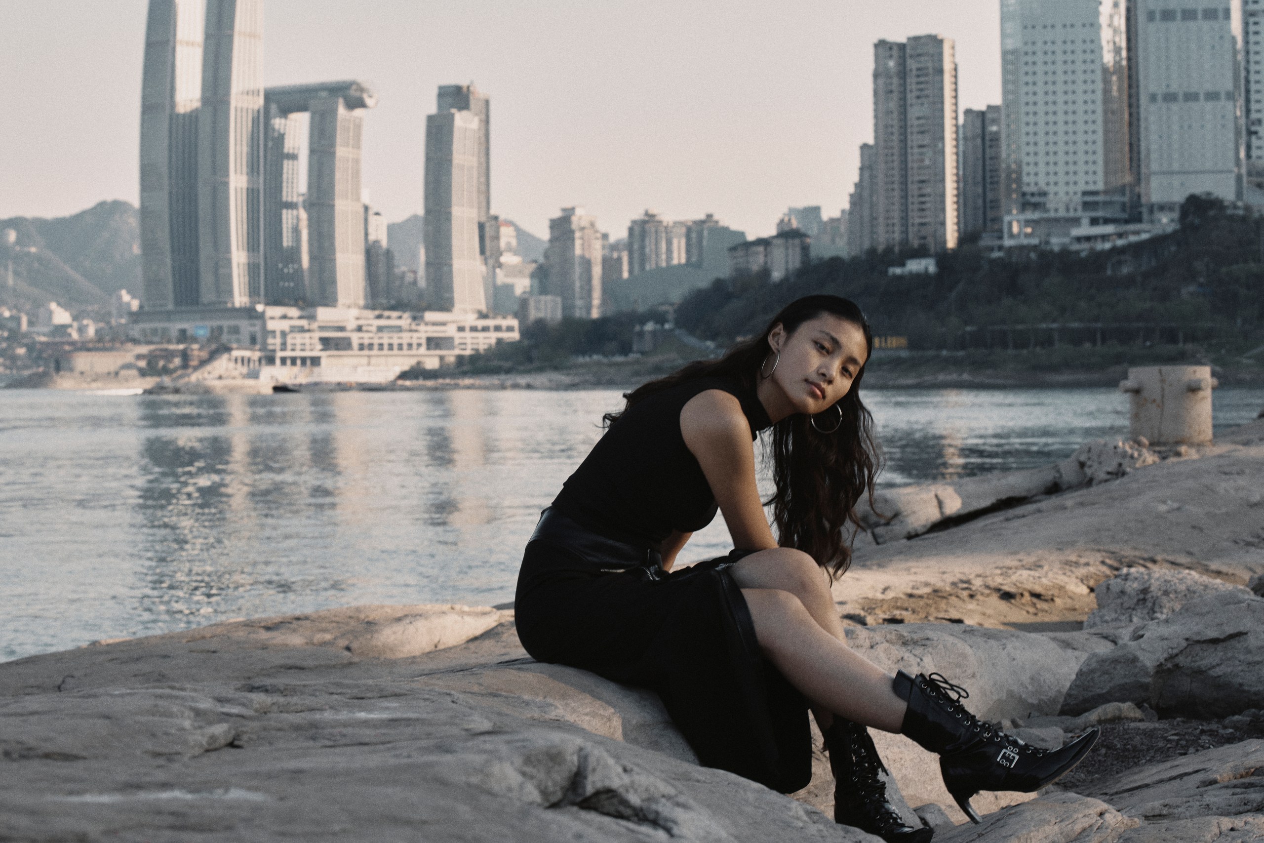 An editorial portrait on the rocky riverside in Chongqing, with the city skyline behind, photographed by Hill Xue